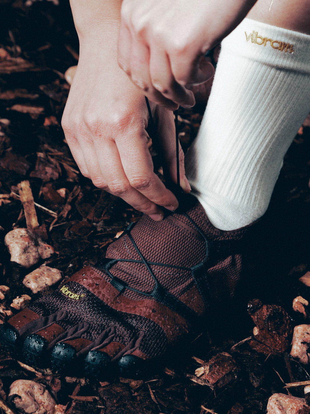 Person tying a brown vibram spidrwalk shoes with Vibram sole on a natural ground surface.